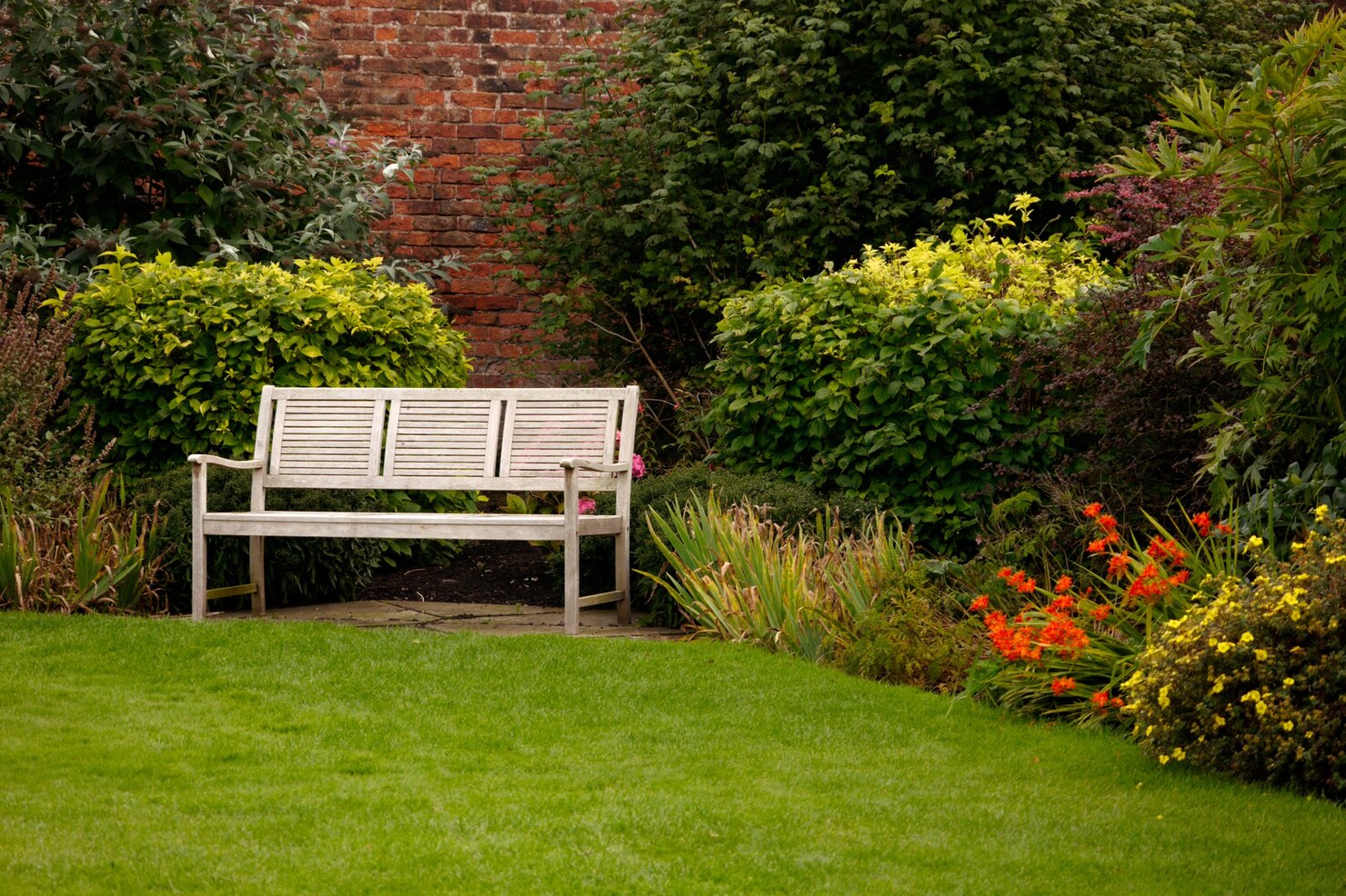 A peaceful UK garden with a wooden bench, lush green lawn, colourful borders and a brick wall